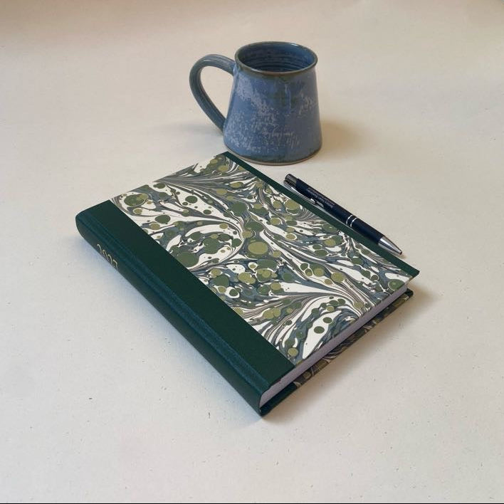 A hardcover notebook with a green and olive green marbled cover lies on a light beige surface next to a black pen, with a blue-gray ceramic mug in the background.