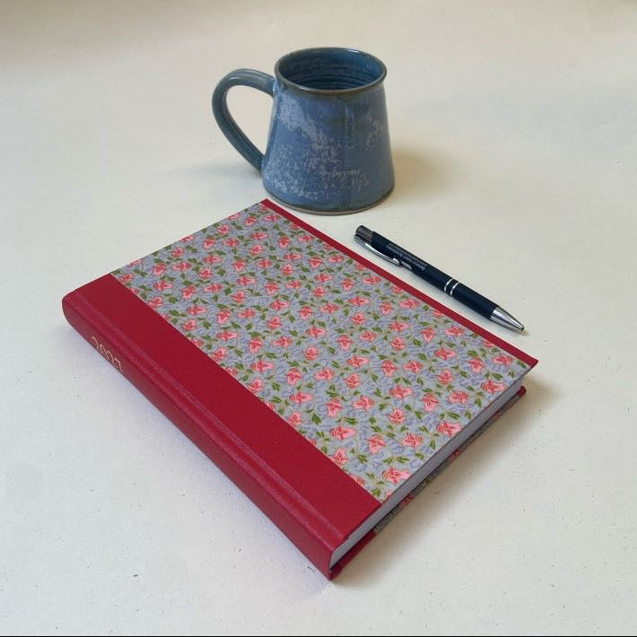 A hardcover notebook with a red spine and a floral-patterned cover lies on a light surface next to a blue ceramic mug and a black pen.