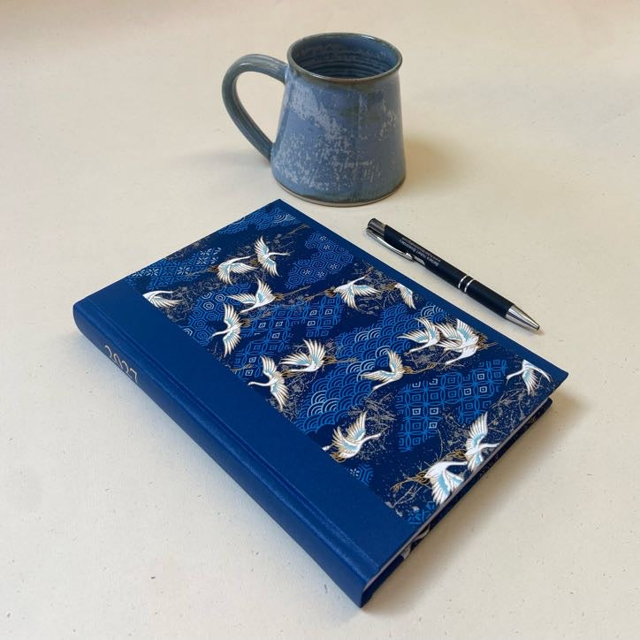 A blue hardcover notebook with a pattern of white cranes and blue wave motifs, styled on a light beige surface next to a black pen and a blue ceramic mug.