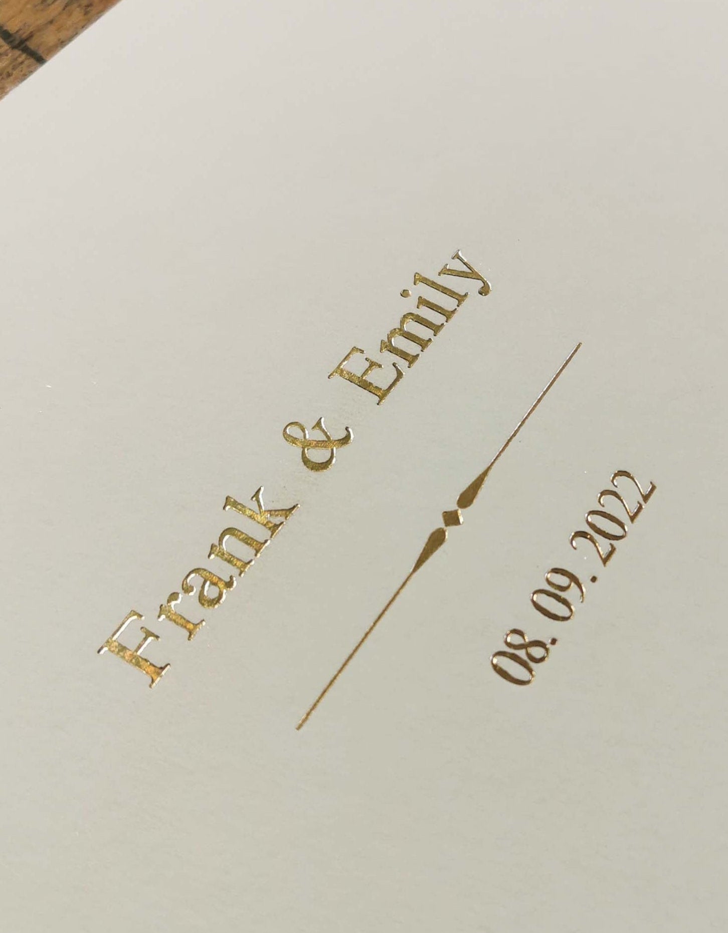 Beige card with gold text 'Frank & Emily' and date '08.09.2023' on a wooden surface