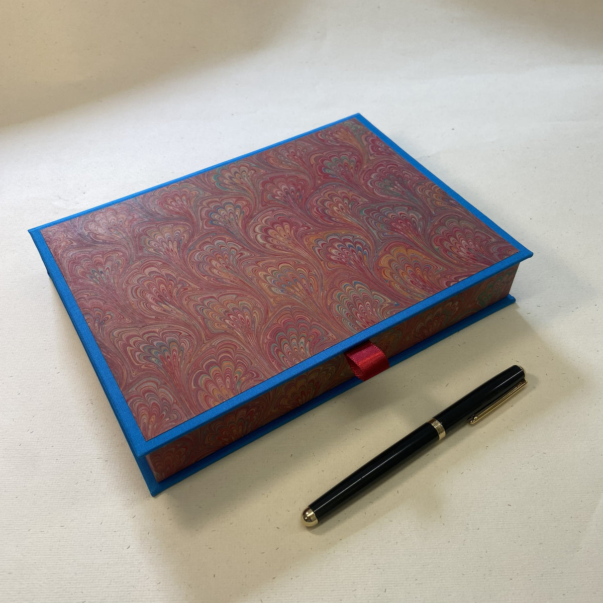 A rectangular marbled paper writing set box with a blue border and a small red pull tab, shown on a light beige surface next to a black pen.