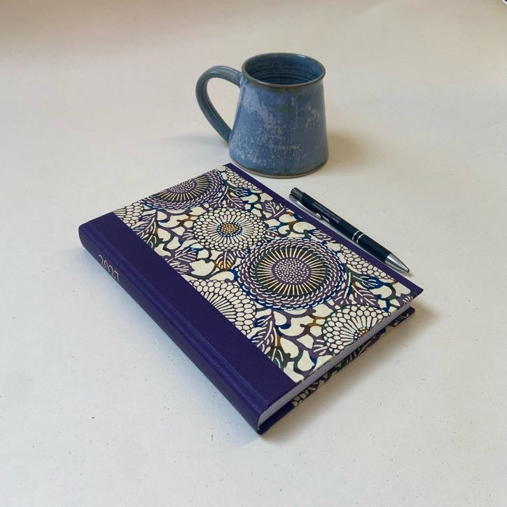 A hardcover notebook with a purple spine and cover, featuring a detailed floral mandala pattern on the front, styled on a light beige surface next to a blue ceramic mug and a black pen.