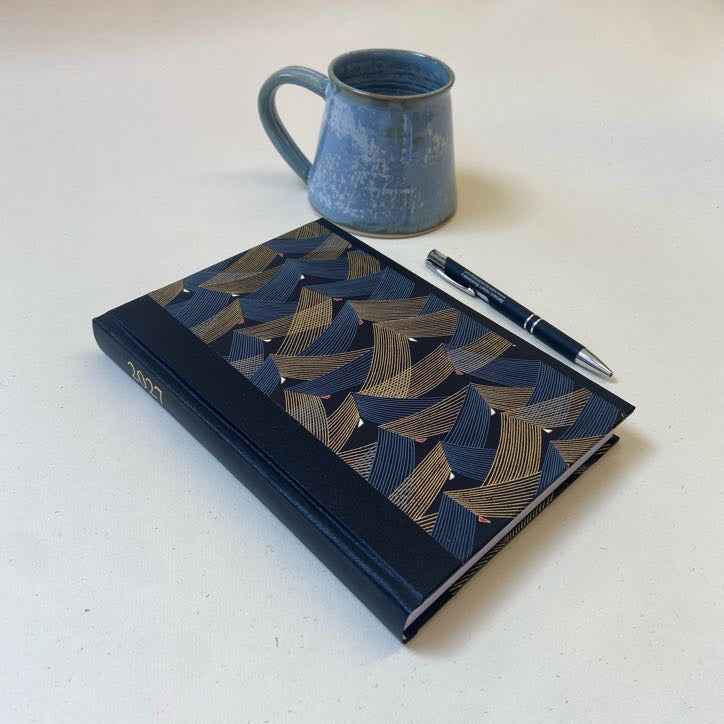 A hardcover notebook with a navy blue and gold geometric patterned cover lies on a light beige surface, styled with a blue ceramic mug and a black pen.
