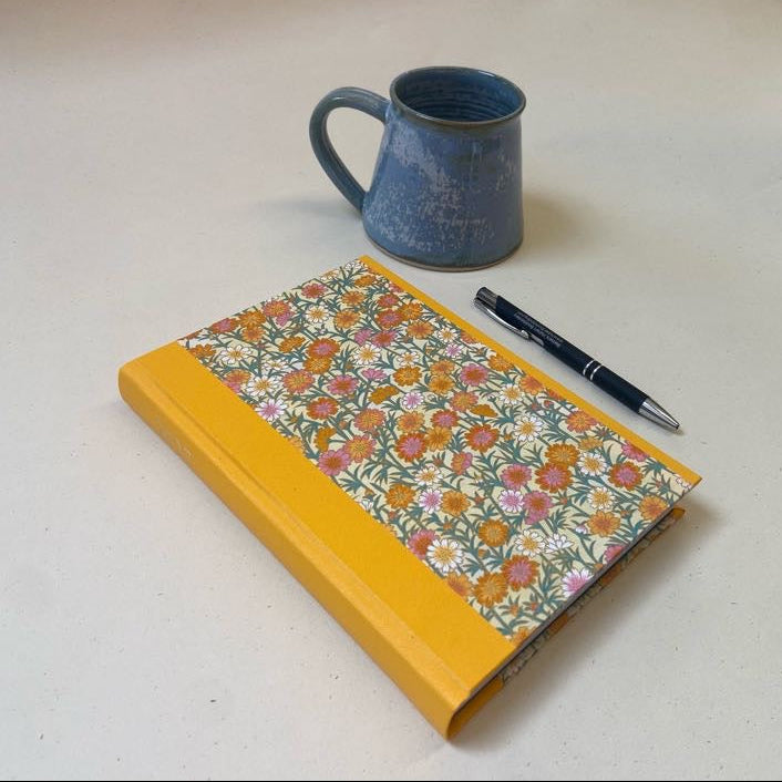 A hardcover notebook with a mustard yellow spine and a floral patterned cover lies on a light beige surface, styled with a blue ceramic mug and a black pen.
