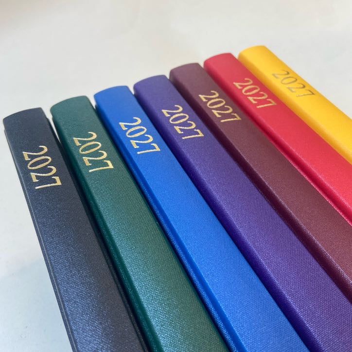 Six hardcover 2027 journals arranged diagonally in a fan shape, showcasing different colors: navy, green, blue, purple, red, and yellow, each with '2027' embossed in gold on the spine.