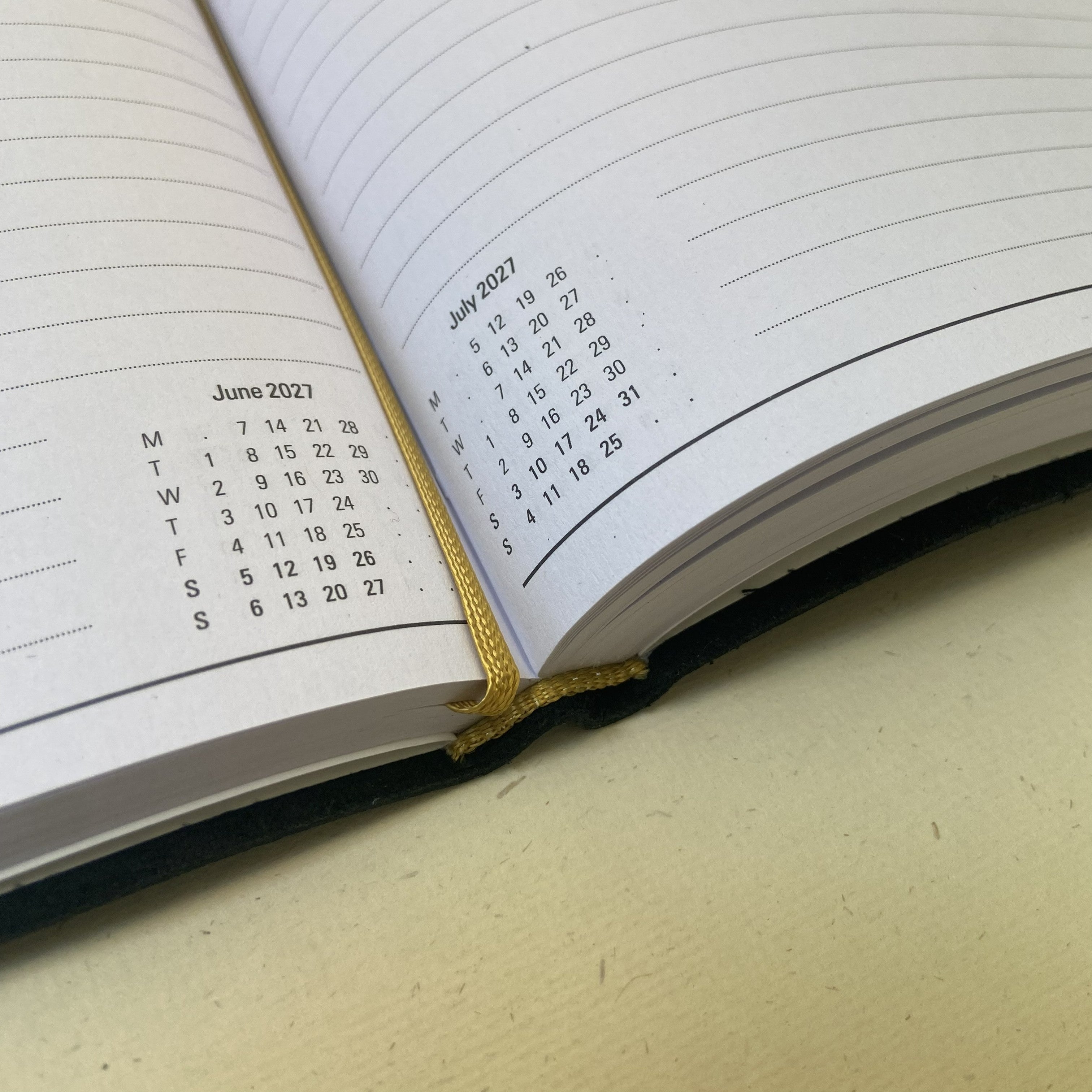 An open lined notebook or planner with a visible June 2027 calendar page on the right and a small monthly calendar on the left, held together by a gold elastic band and a black cover, photographed on a light beige surface.