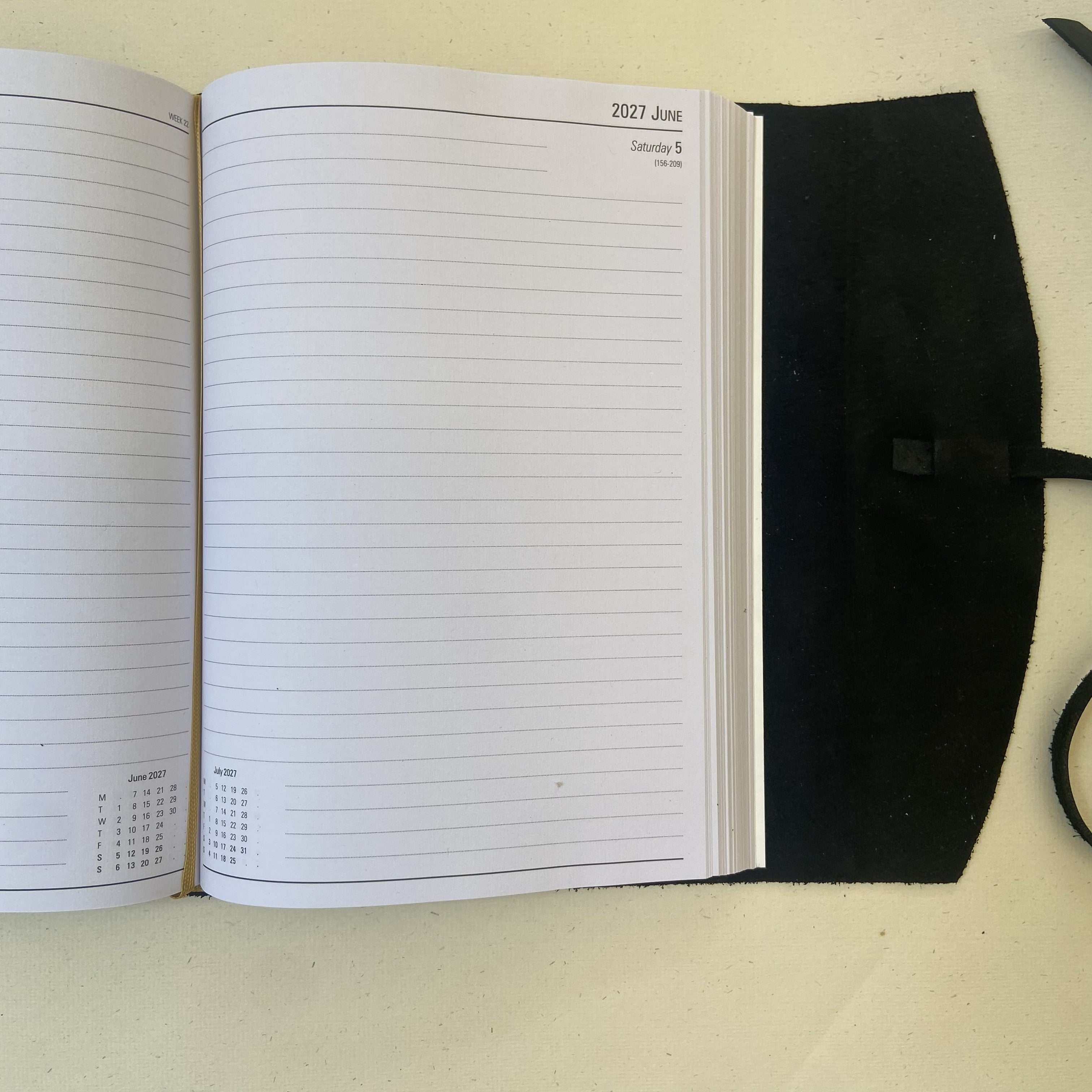 An open lined planner or notebook with a black cover, photographed from above on a light beige surface. The left page shows a calendar spread, and the right page has the date 2027 June and a small calendar inset near the bottom.